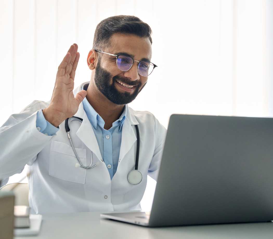 A medical professional in a lab coat is smiling and waving at a laptop camera, likely engaging in a virtual consultation.