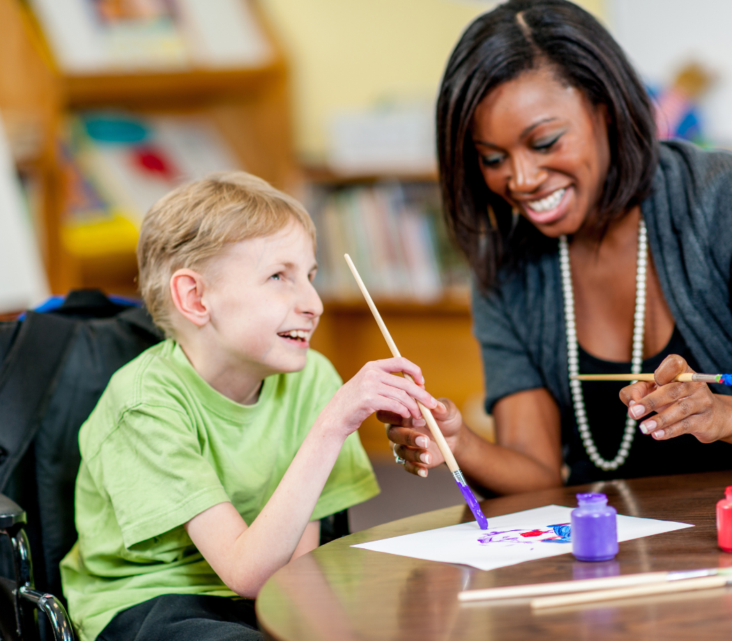 Child with disabilities painting on a paper at a table with adult aiding.